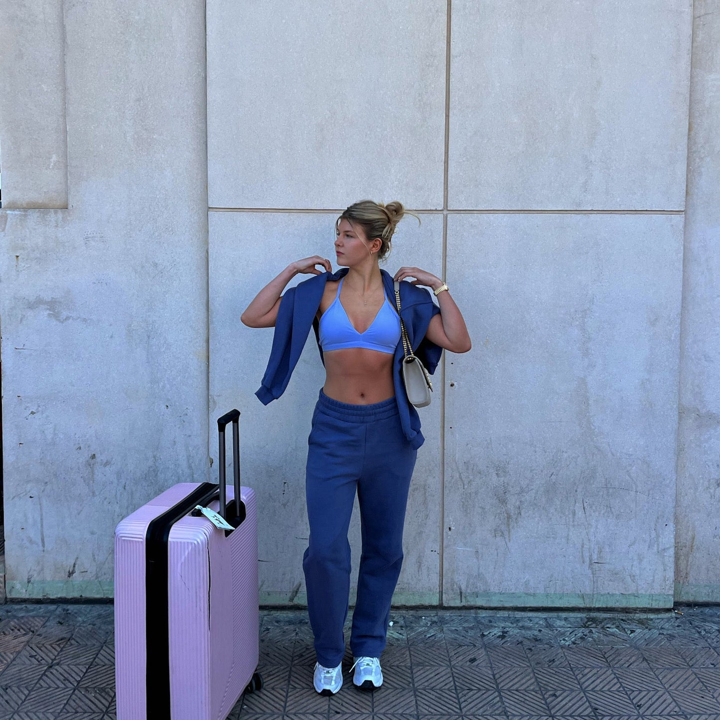Woman in blue sos tracksuit standing next to a pink suitcase against a concrete wall.