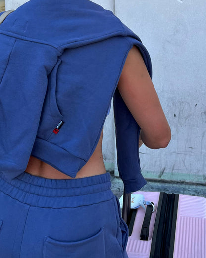 Person wearing a blue sos tracksuit with a visible logo tags, standing against a concrete wall.