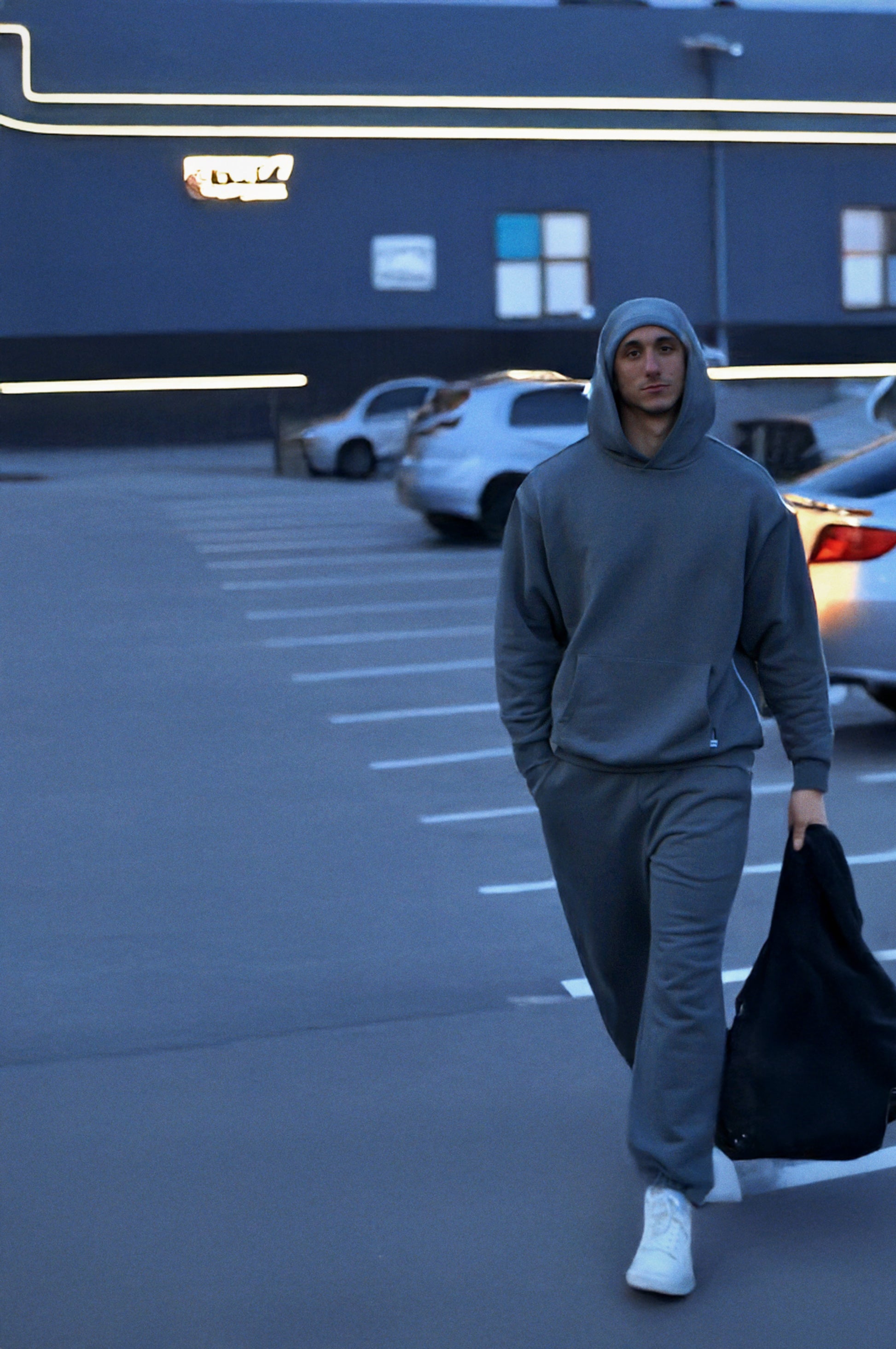 Man in gray sos hoodie and pants walking in a parking lot with cars in the background