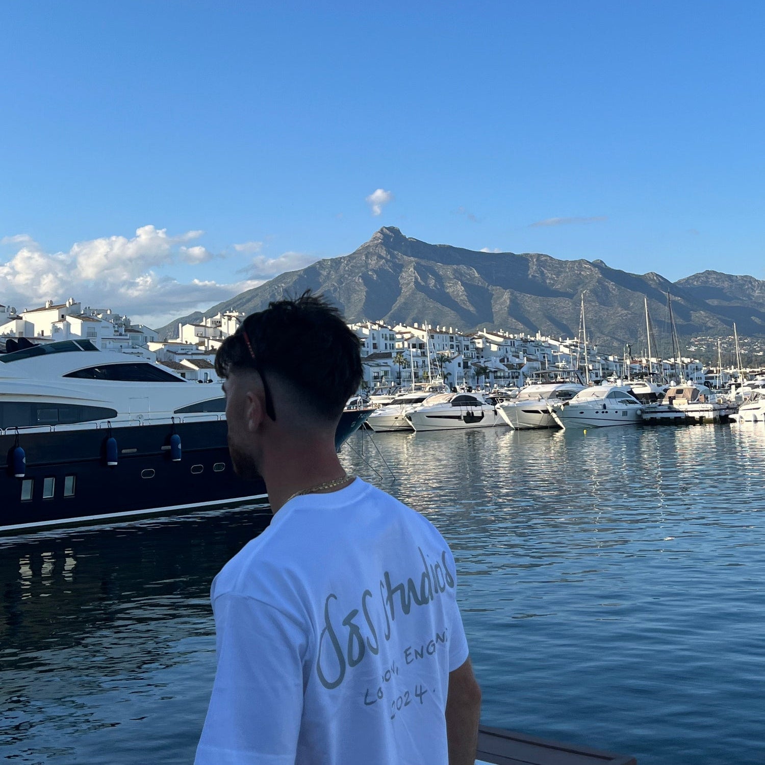 Person standing by a marina with boats and mountains in the background wearing sos studios
