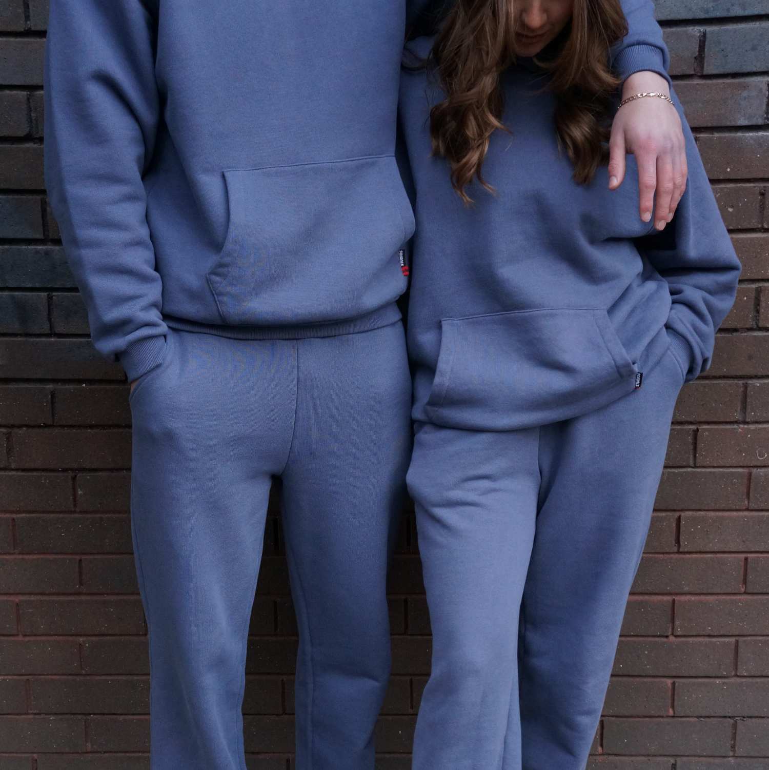 Two people wearing matching blue sos tracksuits standing against a brick wall.