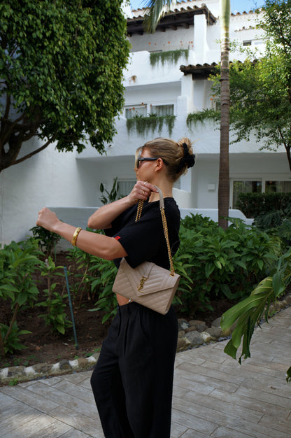 Woman in black outfit with beige bag standing on a sidewalk with greenery and a building in the background